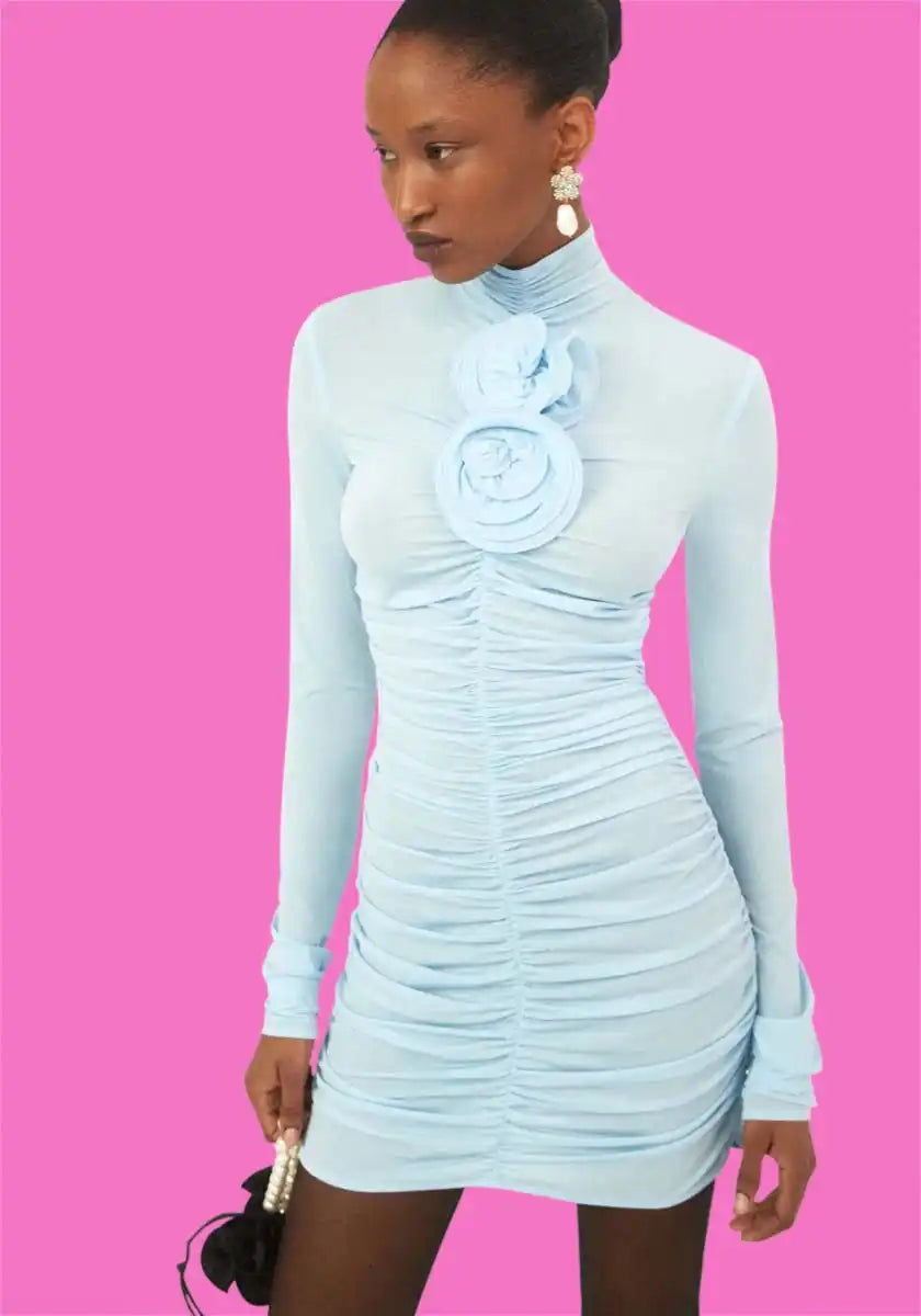 A woman modeling a light blue, long-sleeved, ruched mini dress with a floral appliqué at the neckline, paired with black sheer tights and white pearl drop earrings.